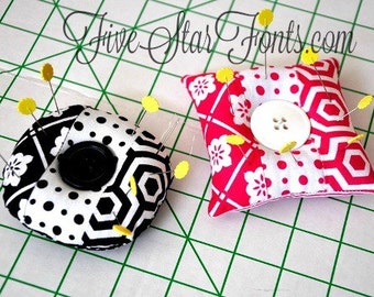 Peut inclure: Deux coussinets à épingles en tissu avec un bouton au centre. Un coussin est noir et blanc avec des motifs géométriques. L'autre coussin est rose et blanc avec un motif floral. Les deux coussinets ont des épingles jaunes qui en sortent.