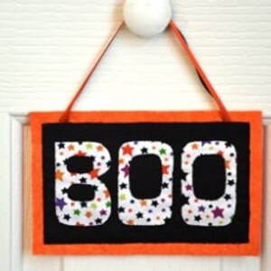 May include: A black felt Halloween door hanger with an orange border. The word "BOO" is written in white letters with a colorful star pattern.