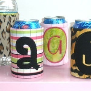 May include: A collection of beverage insulators, including a water bottle sleeve and can coolers. The coolers feature various designs, such as a leopard print, striped pattern, and initial letters. The colors include pink, green, black, and tan.