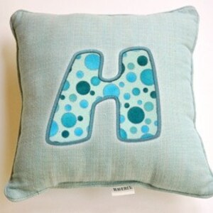 May include: A light blue throw pillow with a large, blue and green polka dot letter "H" in the center.