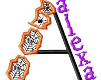 May include: A purple and black embroidered letter A with a spider and spiderweb design. The letter is decorated with three orange spiderwebs with black spiders.