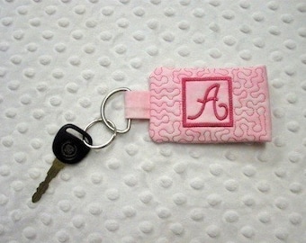 May include: Pink fabric key chain with a pink embroidered letter A in a square frame. The key chain is attached to a silver key ring.
