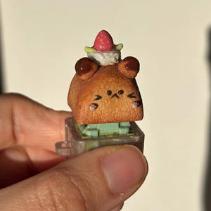 May include: A brown, bear-shaped artisan keycap with dark brown ears and a cute face. The keycap is topped with a strawberry and whipped cream. The keycap is mounted on a clear, rectangular switch.