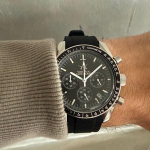 May include: A black and silver wristwatch with a black band. The watch face has multiple sub-dials and the text "von Arx Swiss Made". The watch is on a wrist, with a gray sleeve visible.