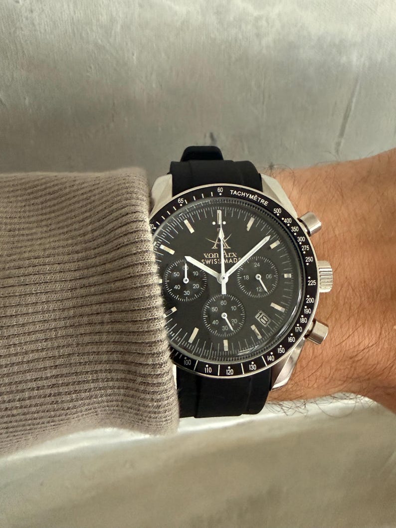 May include: A black and silver wristwatch with a black band. The watch face has multiple sub-dials and the text "von Arx Swiss Made". The watch is on a wrist, with a gray sleeve visible.