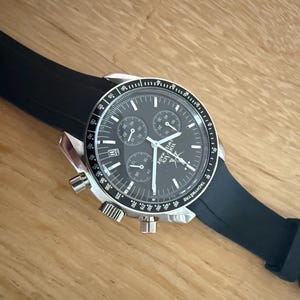 May include: A stainless steel chronograph watch with a black dial and a black rubber strap. The watch face has three sub-dials and a date window. The watch is on a wooden surface.