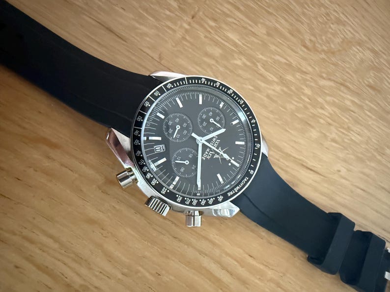 May include: A stainless steel chronograph watch with a black dial and a black rubber strap. The watch face has three sub-dials and a date window. The watch is on a wooden surface.