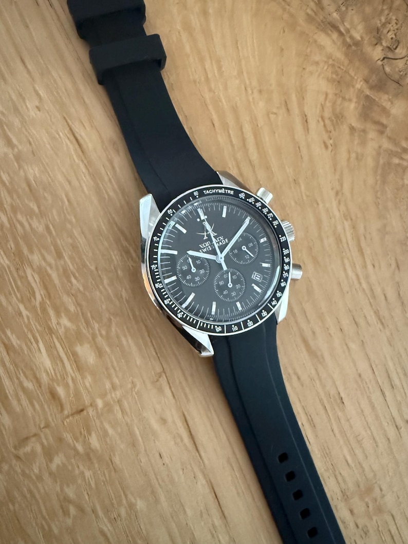 May include: A black wristwatch with a black rubber strap. The watch face is dark gray with silver accents and multiple sub-dials. The bezel has a tachymeter scale. The watch is labeled "SWISS MADE".