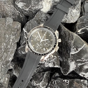 May include: A black and silver wristwatch with a black rubber strap. The watch face is dark with multiple subdials and silver hands. The watch is lying on a bed of black rocks.