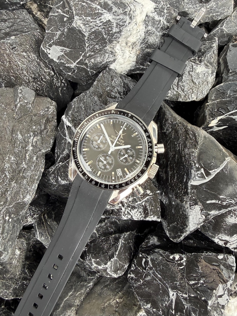 May include: A black and silver wristwatch with a black rubber strap. The watch face is dark with multiple subdials and silver hands. The watch is lying on a bed of black rocks.