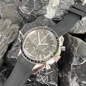 May include: A silver wristwatch with a black face and a black rubber strap. The watch has multiple sub-dials and the text "VO Arx WISS MADE". The watch is set against a background of dark grey rocks.