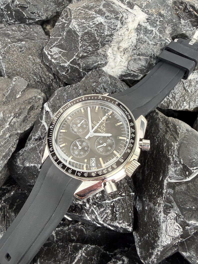 May include: A silver-toned wristwatch with a black dial and a black rubber strap. The watch face features multiple sub-dials and the text "VO Arx WISS MADE". The watch is set against a background of dark gray rocks.