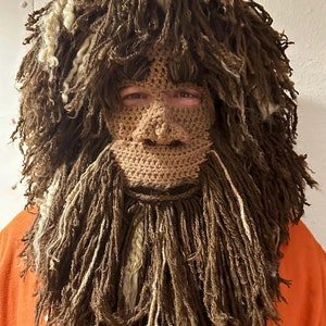 Könnte beinhalten: Eine braune gehäkelte Maske mit langem Bart und Haaren. Die Maske bedeckt das Gesicht der Person von der Stirn bis zum Kinn. Die Maske ist aus Garn gefertigt und hat eine strukturierte Oberfläche.