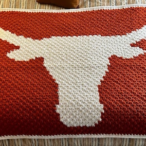 May include: A crocheted throw blanket with a white Longhorn steer head design on an orange background.