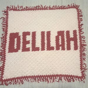 Peut inclure: Une couverture crochetée blanche et rose avec le nom "DELILAH" en grandes lettres roses. La couverture a une surface texturée et une bordure à franges de couleur rose assortie. La couverture est de forme carrée.
