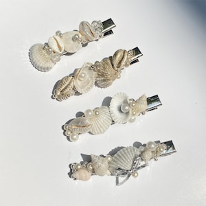 Op de afbeelding: Vier haarclips met zeeschelpen en zilverkleurige metalen sluitingen. Elke clip heeft een unieke opstelling van witte en bruine zeeschelpen, parels en kleine kralen. De clips variëren in schelptypes en decoratieve details.