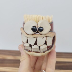May include: A whimsical ceramic toothpick holder with a cartoonish face. The holder features large, white teeth, big round eyes with black pupils, and a wide, smiling mouth. The holder is filled with a bundle of toothpicks.
