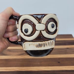May include: A brown ceramic mug with a whimsical owl face design. The mug features large, round eyes with black pupils, a white beak, and a textured, stylized mouth. The handle and the base of the mug are dark brown.