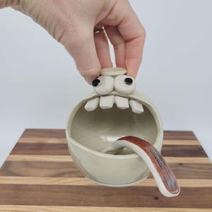 May include: A whimsical, off-white ceramic bowl with a cartoonish face. The bowl features large, black-eyed eyeballs, a row of teeth, and a tongue with red and brown markings. The bowl is held by a hand.