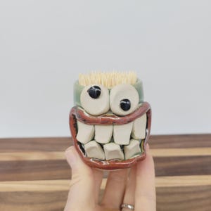 May include: A whimsical ceramic toothpick holder shaped like a grinning face. The holder features large, cartoonish eyes, a wide red mouth with prominent white teeth, and a green rim. The holder is filled with wooden toothpicks.