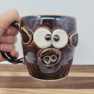May include: A brown ceramic mug with a pig face design. The mug features large white eyes, a snout, and ears. The handle is a twisted design. The mug is on a wooden surface.