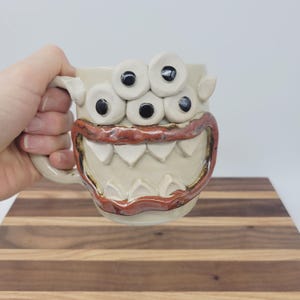 May include: A beige ceramic mug with a monster face design. The mug features five white, round eyes with black pupils, a red and brown mouth, and sharp white teeth. The mug is held by a hand.