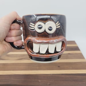 May include: A brown ceramic mug with a whimsical face design. The mug features large white teeth, wide eyes, and a smiling mouth. The mug is held by a hand, and the background is a wooden cutting board.