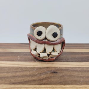 May include: A ceramic mug with a whimsical design. The mug features a wide, toothy grin, large white eyes with black pupils, and a brown rim. The mug is a unique, handmade piece.