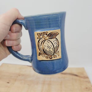 May include: A blue ceramic mug with a brown square design featuring a peach and the text "Just Peachy".
