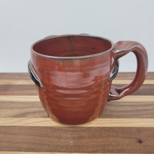 May include: A handmade ceramic mug with a rich, reddish-brown glaze. The mug features a unique handle and decorative accents on the sides. The interior shows darker tones. The mug is designed for hot beverages.