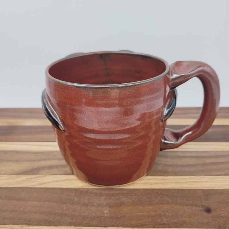 May include: A handmade ceramic mug with a rich, reddish-brown glaze. The mug features a unique handle and decorative accents on the sides. The interior shows darker tones. The mug is designed for hot beverages.