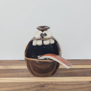 May include: A whimsical, brown ceramic bowl with a cartoonish face design. The bowl features large white teeth, bulging eyes, and a tongue-shaped spoon. The interior is dark brown, and the exterior has a textured finish.
