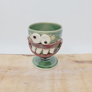 May include: A whimsical, handmade ceramic goblet with a green glaze. The goblet features a cartoonish face with large white eyes, a red mouth, and white teeth. The base and interior are also green, creating a unique, playful design.