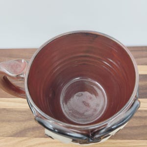 May include: A handmade ceramic mug with a wide, cylindrical shape. The interior is a rich, reddish-brown color, contrasting with the lighter exterior. The mug features a sturdy handle and a decorative design near the base, perfect for enjoying hot beverages.