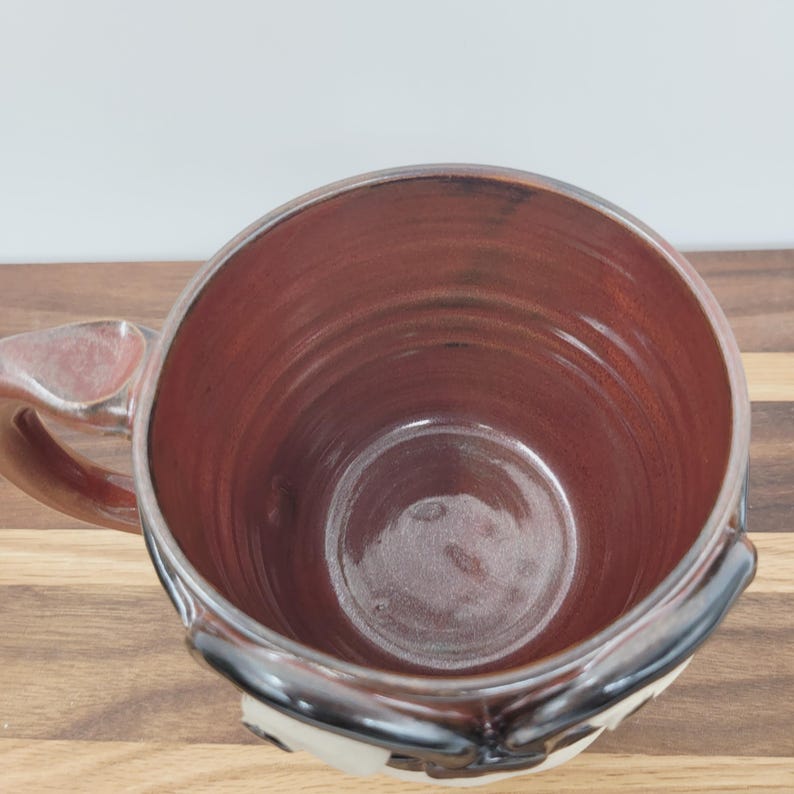 May include: A handmade ceramic mug with a wide, cylindrical shape. The interior is a rich, reddish-brown color, contrasting with the lighter exterior. The mug features a sturdy handle and a decorative design near the base, perfect for enjoying hot beverages.