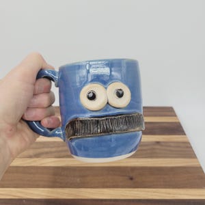 May include: A blue ceramic mug with a whimsical face design. The mug features large, round eyes and a textured, dark brown mustache. The mug is held by a hand, showcasing its handle. The mug is on a wooden surface.