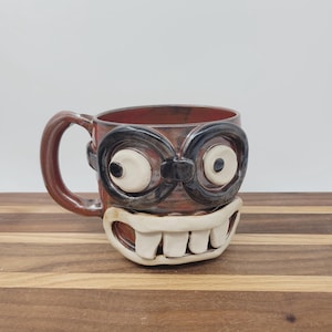 May include: A handmade ceramic mug with a whimsical face design. The mug features large, cartoonish eyes with black frames and a wide, toothy grin. The mug is brown with a red interior and a brown handle.