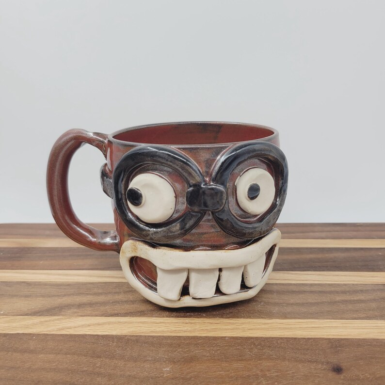 May include: A handmade ceramic mug with a whimsical face design. The mug features large, cartoonish eyes with black frames and a wide, toothy grin. The mug is brown with a red interior and a brown handle.