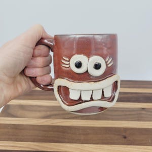 May include: A brown ceramic mug with a whimsical face design. The mug features large, round eyes with black pupils, a wide, toothy grin, and a handle. The mug is held by a hand.