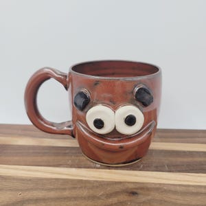 May include: A handmade ceramic mug with a whimsical face design. The mug is primarily brown with black and white accents for the eyes and a smiling mouth. It has a large handle and is suitable for hot or cold beverages.