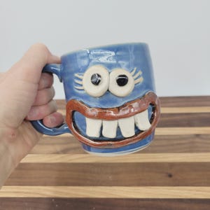 May include: A blue ceramic mug with a whimsical face design. The mug features large, googly eyes, a wide, toothy grin, and a handle. The mug is held in a hand, and the background is a wooden surface.