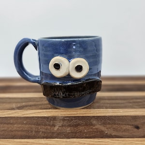 May include: A blue ceramic mug featuring two large, white, circular eyes with black pupils and a wide, dark brown, rectangular mustache attached to the front. The mug has a curved handle on the left side and a smooth, glossy finish throughout.