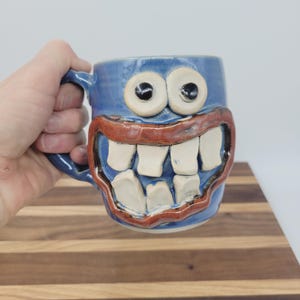 May include: A blue ceramic mug with a cartoonish face design. The mug features large white teeth, a red mouth, and two large, round eyes. The mug is held by a hand, showcasing the handle and overall shape.