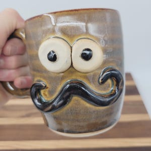 May include: A handmade ceramic mug with a whimsical design. The mug features large, cartoonish eyes and a prominent black mustache. The mug has a brown and tan glaze with a handle on the side.