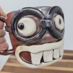 May include: A handmade ceramic mug with a whimsical monster face design. The mug features large, round eyes with black rims, a wide grin with prominent teeth, and a reddish-brown body. The handle is brown.