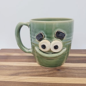 May include: A handmade ceramic mug in a light green color, featuring a whimsical face design. The mug has large, round eyes with black pupils, and a smiling mouth. The handle is curved, and the mug sits on a wooden surface.