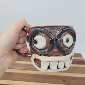 May include: A handmade ceramic mug with a cartoonish face design. The mug features large, white eyes with black outlines, a wide grin with large white teeth, and a brown and black color scheme. The mug is held by a person.