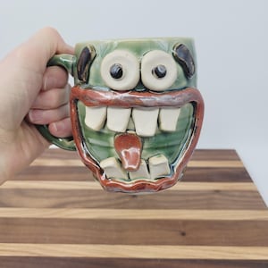 May include: A ceramic mug with a green glaze, featuring a monster face design. The mug has large white teeth, a red tongue, and large, round eyes. The handle is green.
