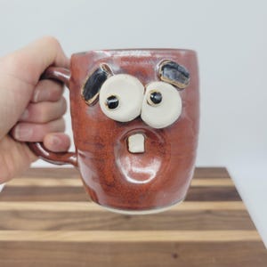 May include: A handmade ceramic mug with a surprised face design. The mug is a reddish-brown color with large white eyes, black accents, and a single tooth. The mug has a handle and is held by a person.