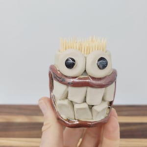 May include: A whimsical, handmade ceramic toothpick holder. The holder is shaped like a grinning face with large, black eyes, a brown mouth, and a set of white teeth. The holder is filled with wooden toothpicks.
